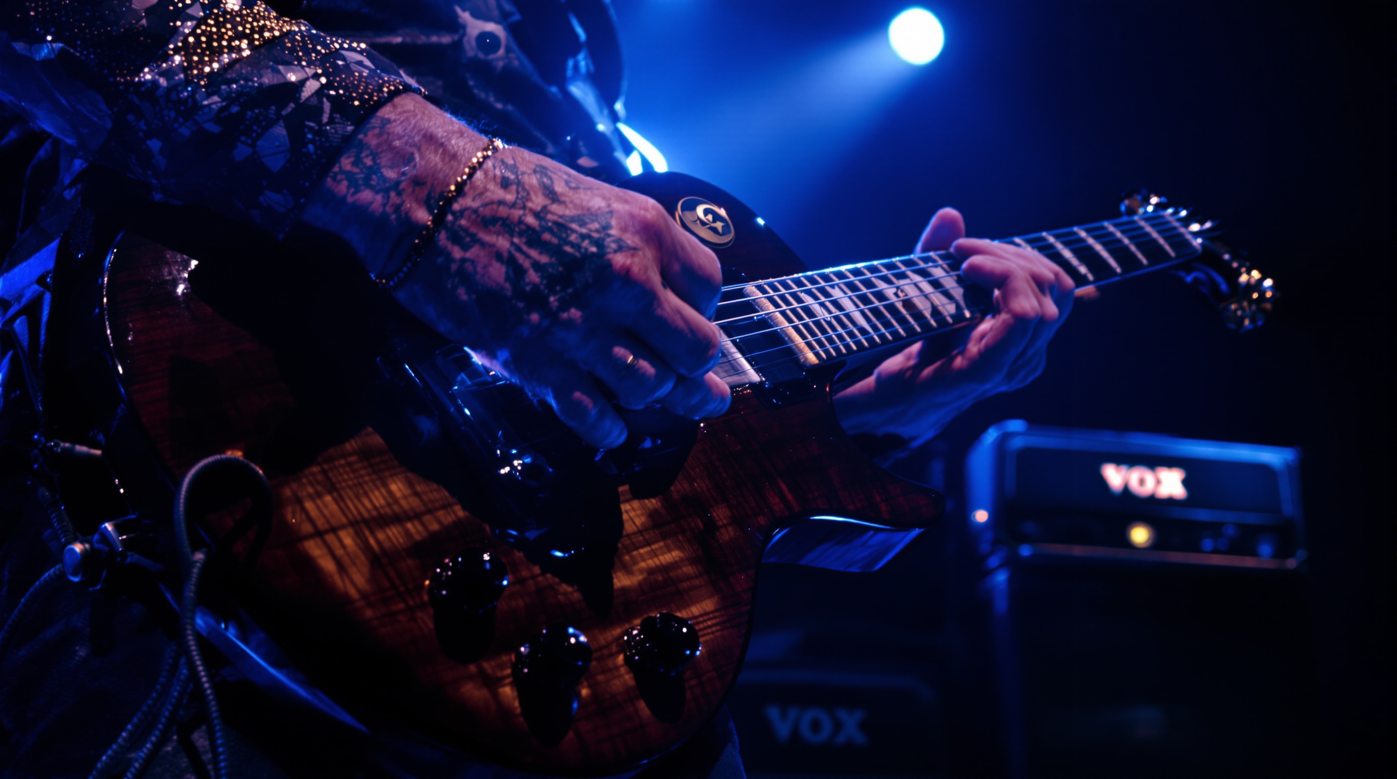 Steve Lukather en concert avec sa guitare emblématique sous une lumière bleutée, amplis Vox-AC-30 visibles en fond.